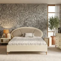 تخت خواب نیلماه با تاج منحنی و رنگ کرم در اتاقی با دیوار طرح خطاطی، آینه آرایشی و گلدان زیتون.